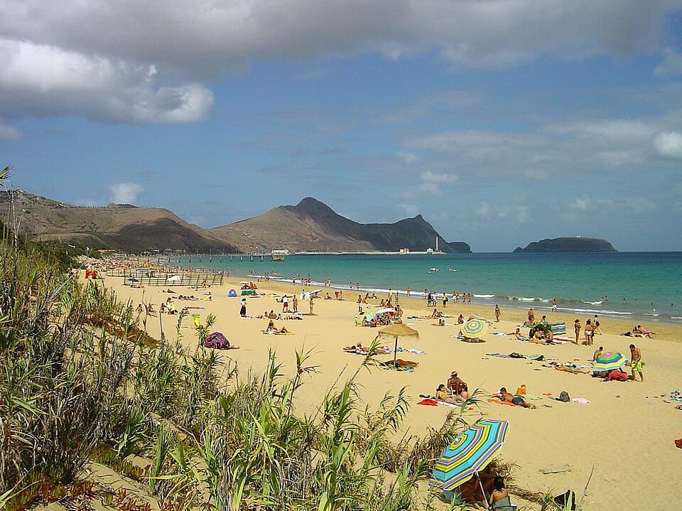 Praia dourada e mar azul do Porto Santo com o relevo da ilha em fundo